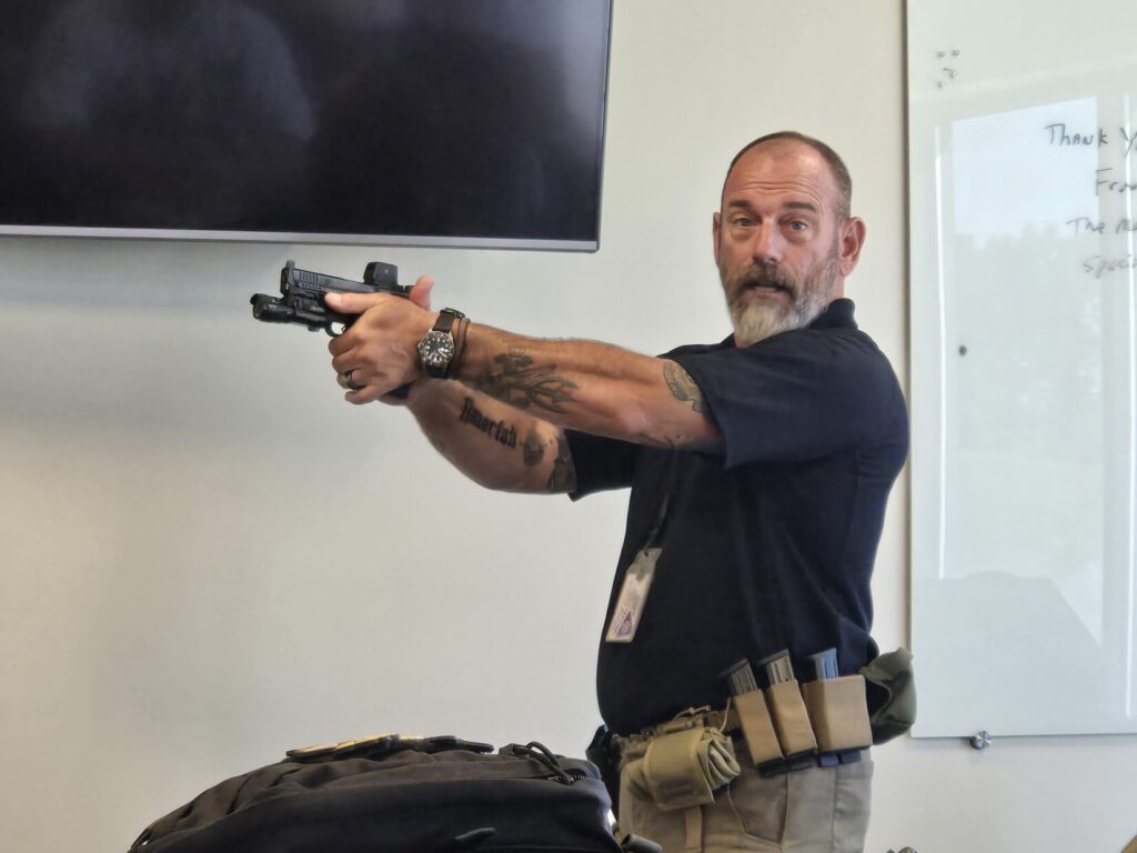A shooting instructor stands in front of a class wiht a black shirt and pistol belt while demonstrating the correct pistol grip.