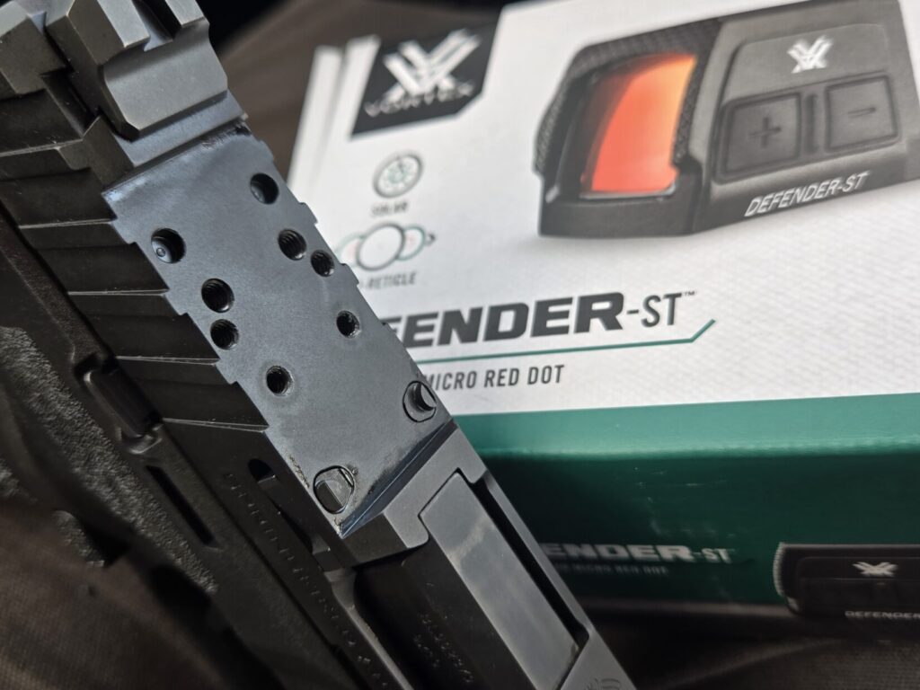 A pistol slide with boxed red dot in the background