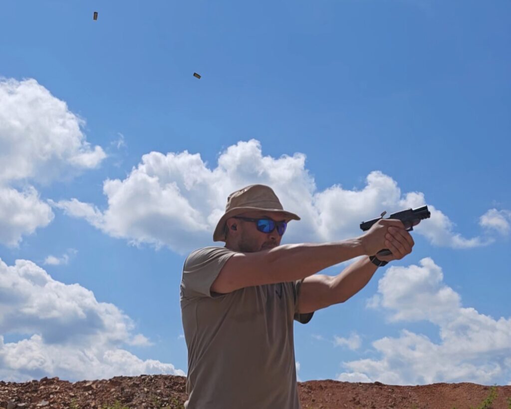 A man with a pistol and three spent casings visible against the sky backdrop