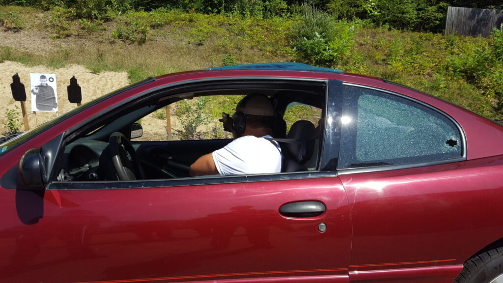 A man in the drivers seat of a car shooting out the passenger side window at a paper target