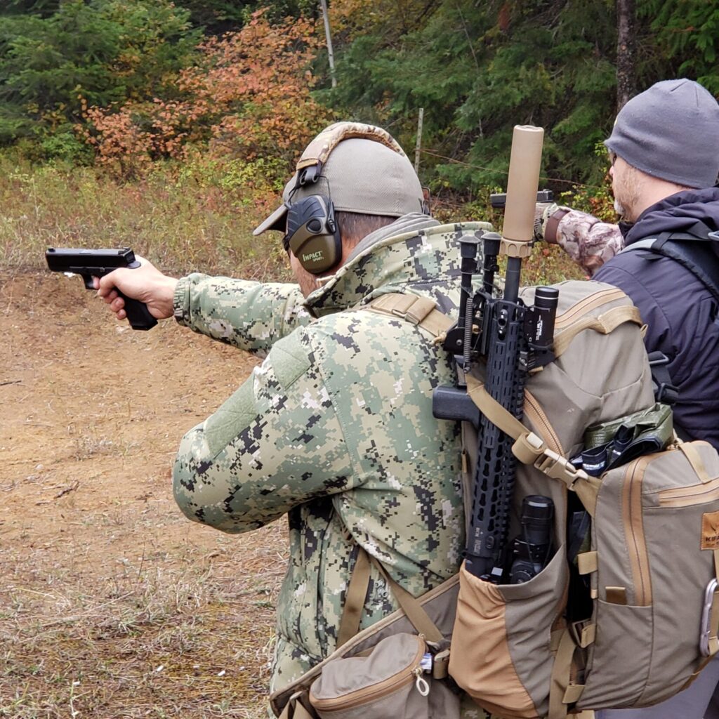 A man in a camo jacket with a backpack and broken down rifle points his pistol