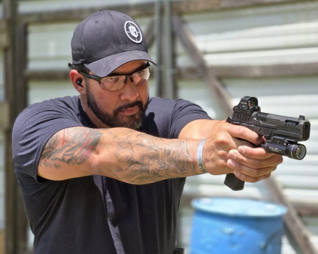 A man in a black shirt and hat holds a pistol with a red dot and light