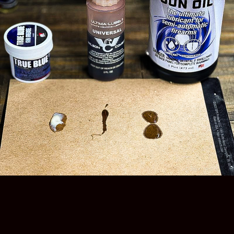 A brown piece of carboard with a blob of white grease on the left, and two other areas with gun oil placed on it.