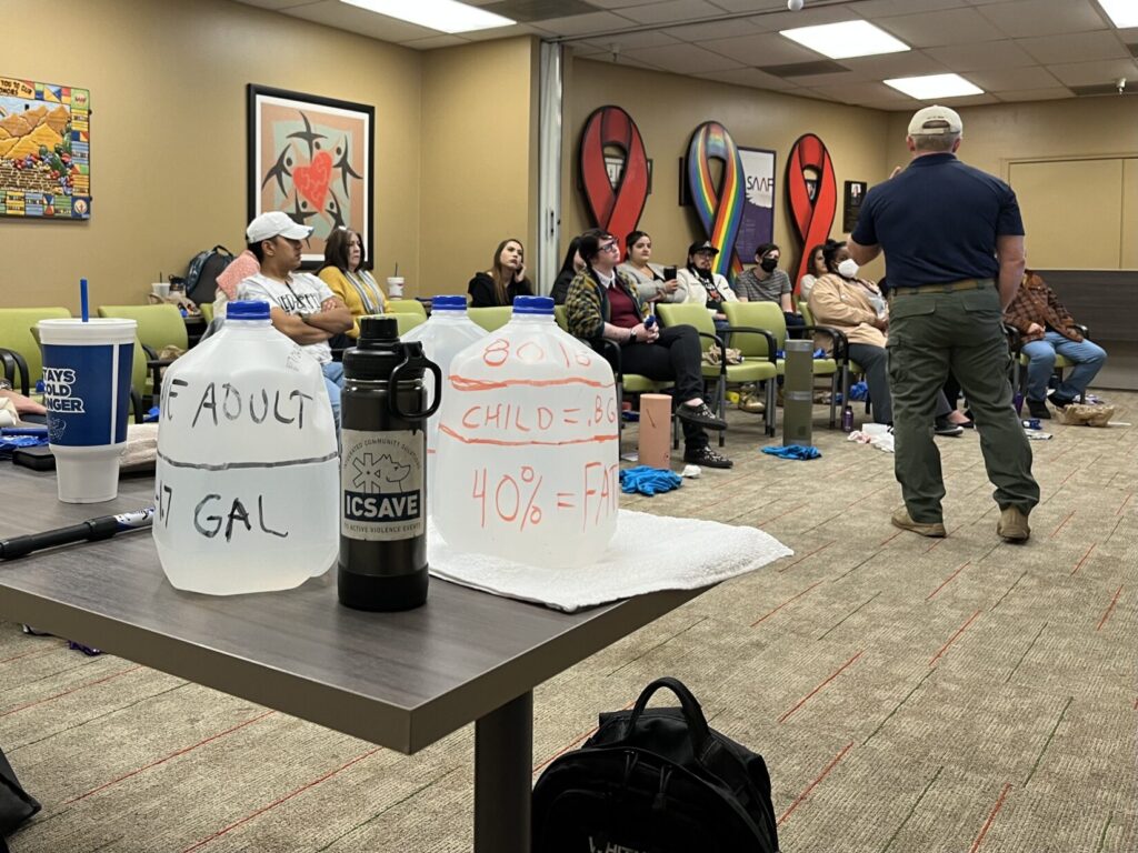 Image of a conference room. An instructor in a blue polo shirt faces the audience. In fthe foreground are three 1-gallon water jugs, marked with lines around them for a demonstration.