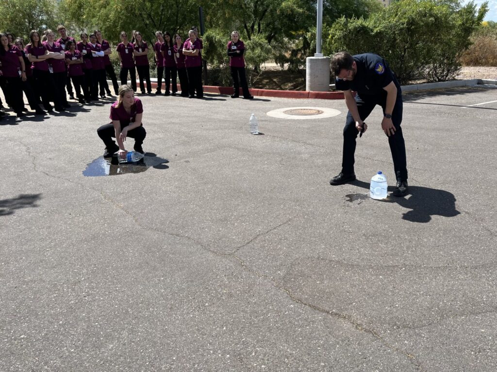 An instructor in a parking lot holds a knife. He's just poked a hole in the bottom of a jug that had been 80% full of water. A nursing student squats down a few feet to the left, holding pressure on a different water jug that had started full. There is a mall puddle of water at her feet.