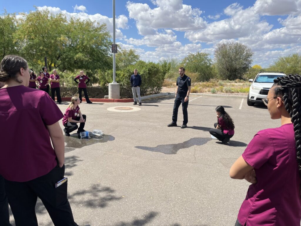 Nursing students squat down next to two 1-gallon jugs of water. Both are leaking. The jug on the right started with less in it and the puddle is smaller.