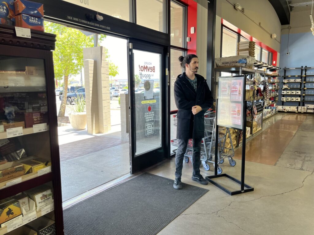 Photo of a man in a heavy coat standing just inside the door of a liquor store. His right hand is inside the coat. The trees outside have green summertime leaves.