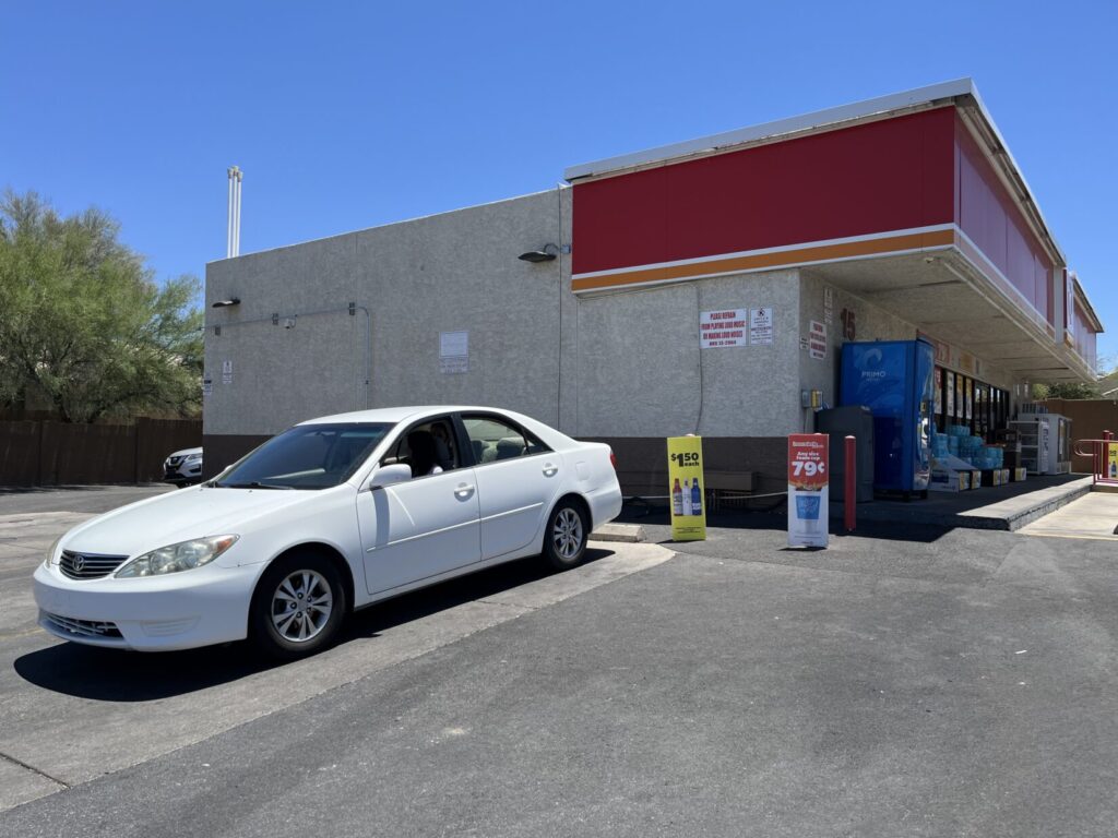 Image of a white Toyota Camry backed into a parking space around the side of a convenience store, out of sight from the windows. Three people are sitting in the car, talking.