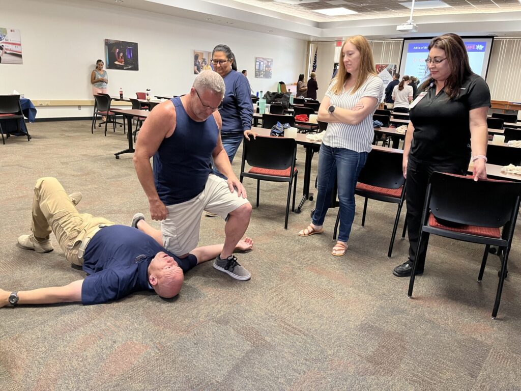 Image of an instuctor in a blue polo shirt lying face up on the floor. A student in a tank top kneels on the instructor's shoulder, as if applying direct pressure to a wound.