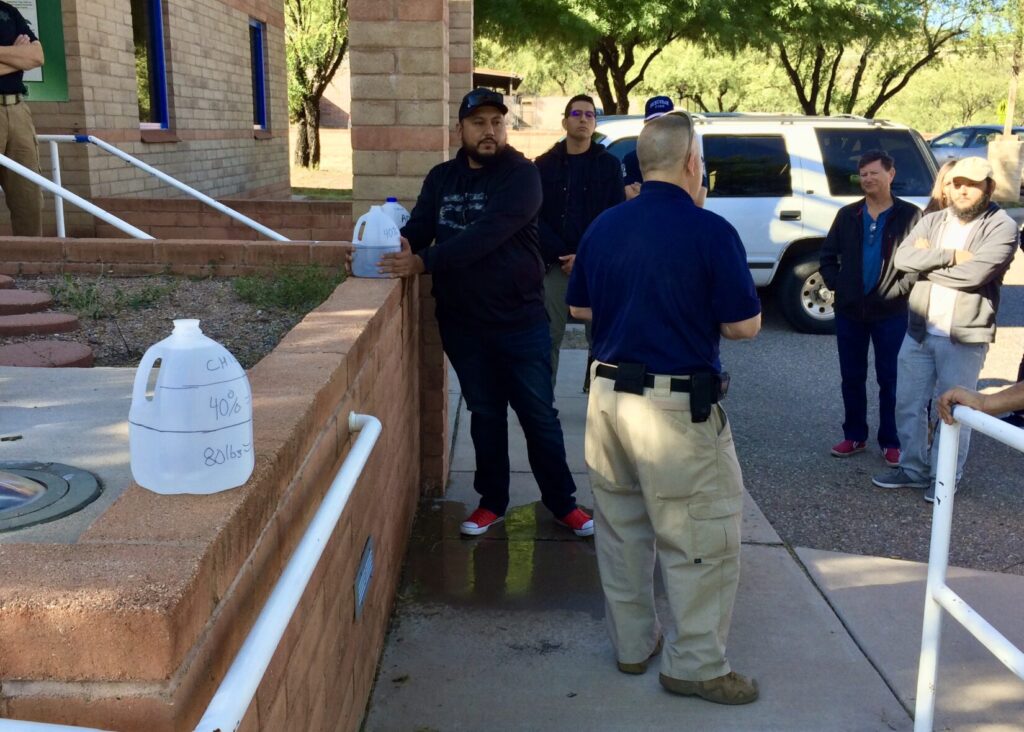 A Bleeding Control class. There is a chest-high brick wall on the left side of the image. 1-gallon water jugs sit atop the wall. The jug in the foreground has two lines around it, but it is full. One of the distant jugs is half-full. The other has been unctured and leaked onto the ground below. A student holds pressure against the hole.
