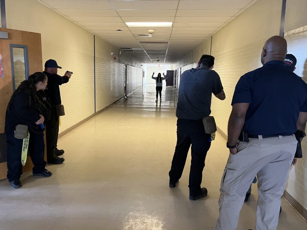 Image of a school hallway. Firefighters learning how police respond to active shooters are keeping role players at the far end of the hall, fleeing simulated active violence, covered with replica firearms.