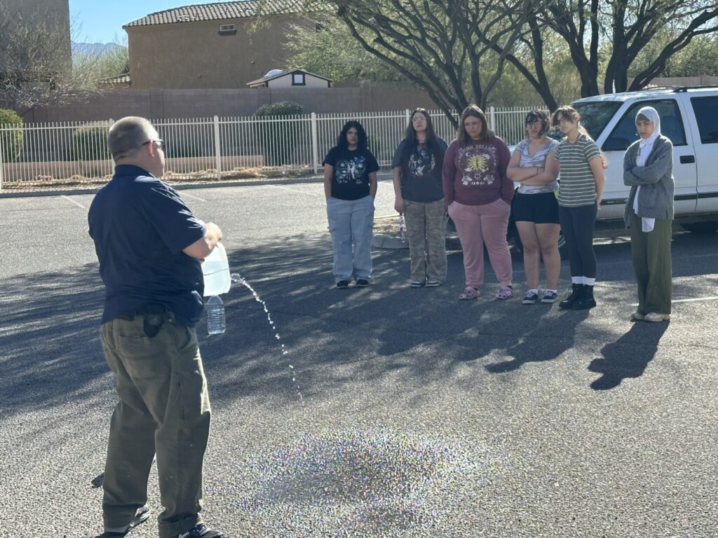 Image of an instructor in a blue Polo holding a 1-gallon water jug in front of him. Water is shooting from a hole in the jug onto the pavement. Several Health Sciences students observe.