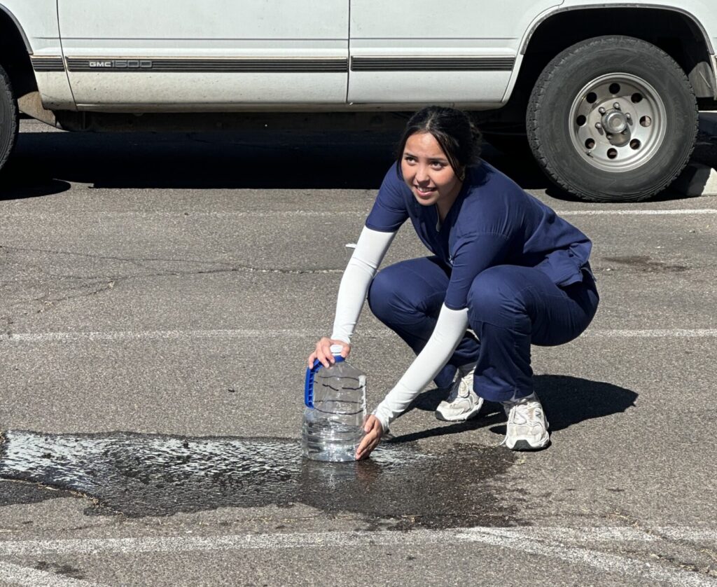 A student in scrubs applies pressure against a puncture in a 1-gallon water jug. There is a large puddle flowing off camera to the left.