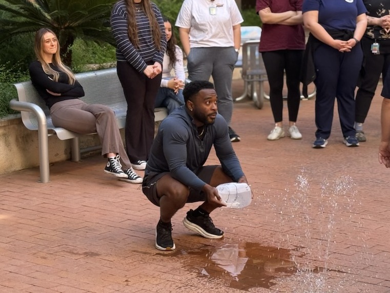 Image of a nursing student squatting down over a brick surface, holding a 1-gallon water jug. There is a water puddle in front of him.