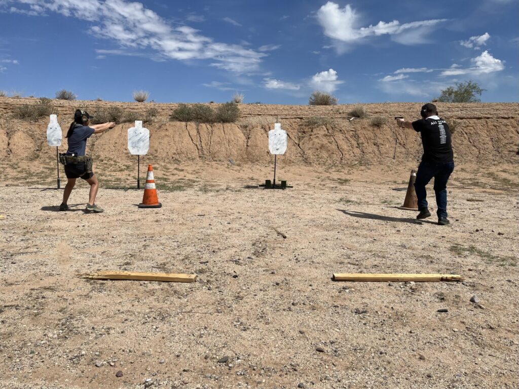 Image of two shooters on a firing range. They are spread out and pointing pistols at a target that is in the 90 degree apex of a right triangle formed by the shooters and the target.