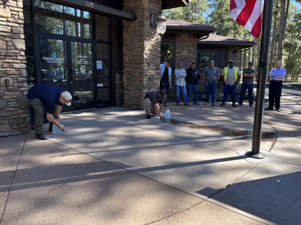 Image of a shaded, sun-dappled walkway in front of a small town municipal building in Arizona's Rim country. A Bleeding Control student kneels on the right, holding pressure over a hole in a leaking 1-gallon jug, while an instuctor on the left punctures a hole in another jug.