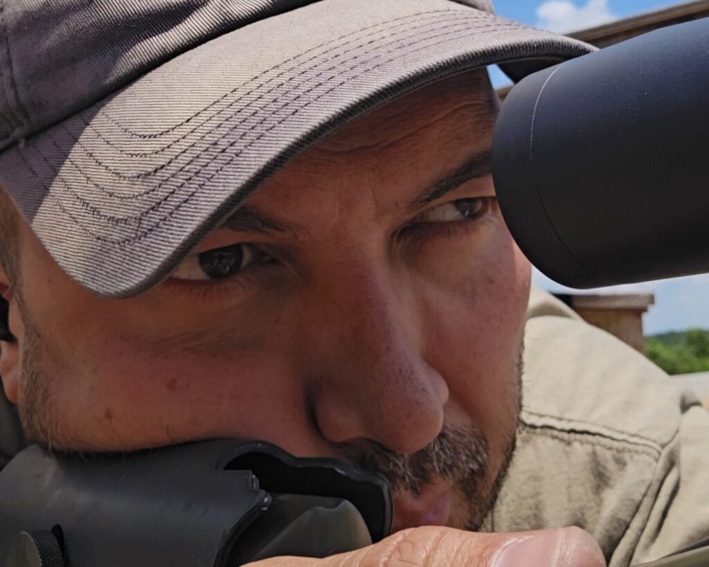 A man resting his face against a rifle stock looking through the optic.
