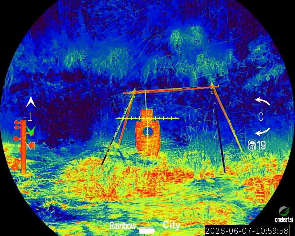 Thermal optic view in "Rainbow" mode and "City" environment. A shooting range setup features a bright orange torso-shaped target hanging from a frame, centered in the yellow crosshairs.