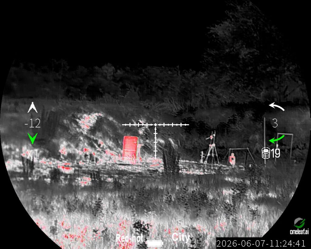 Thermal optic view in "Red-hot" mode and "City" environment. A high-contrast grayscale landscape features a bright red, rectangular structure standing out to the left of the white crosshairs.