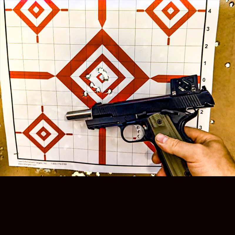 A person holding up a black 1911 pistol against a white and red paper target showing bullet holes in the middle. 