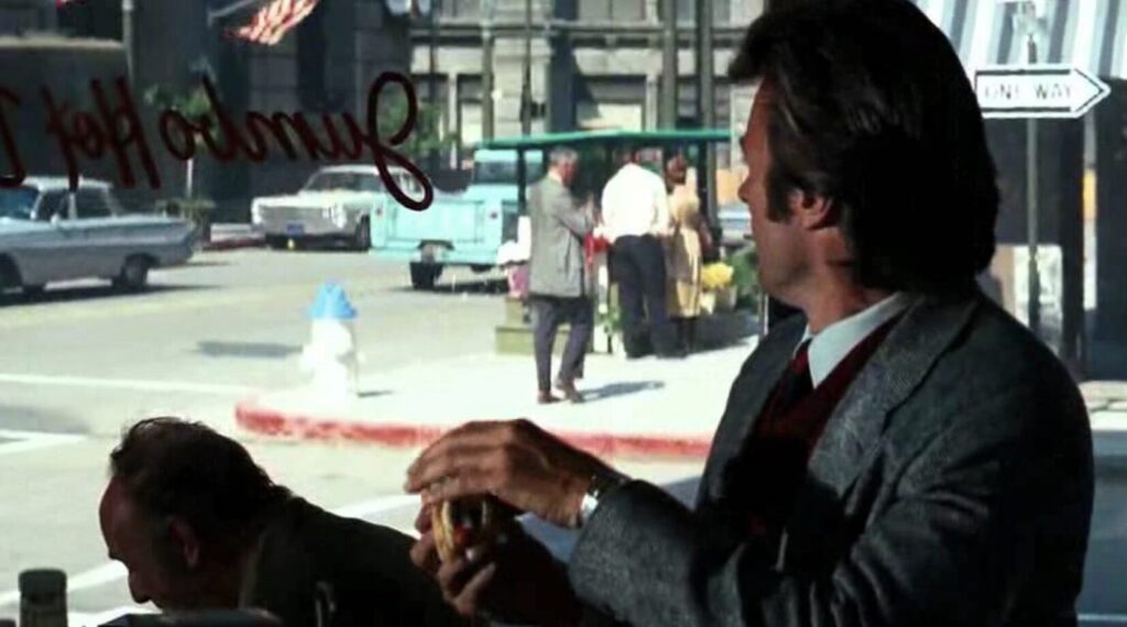 Image of Clint Eastwood as "Dirty Harry" Callahan looking over his shoulder through a plate glass window at a late 1960s or early 1970s tan Ford sedan parked in front of a bank