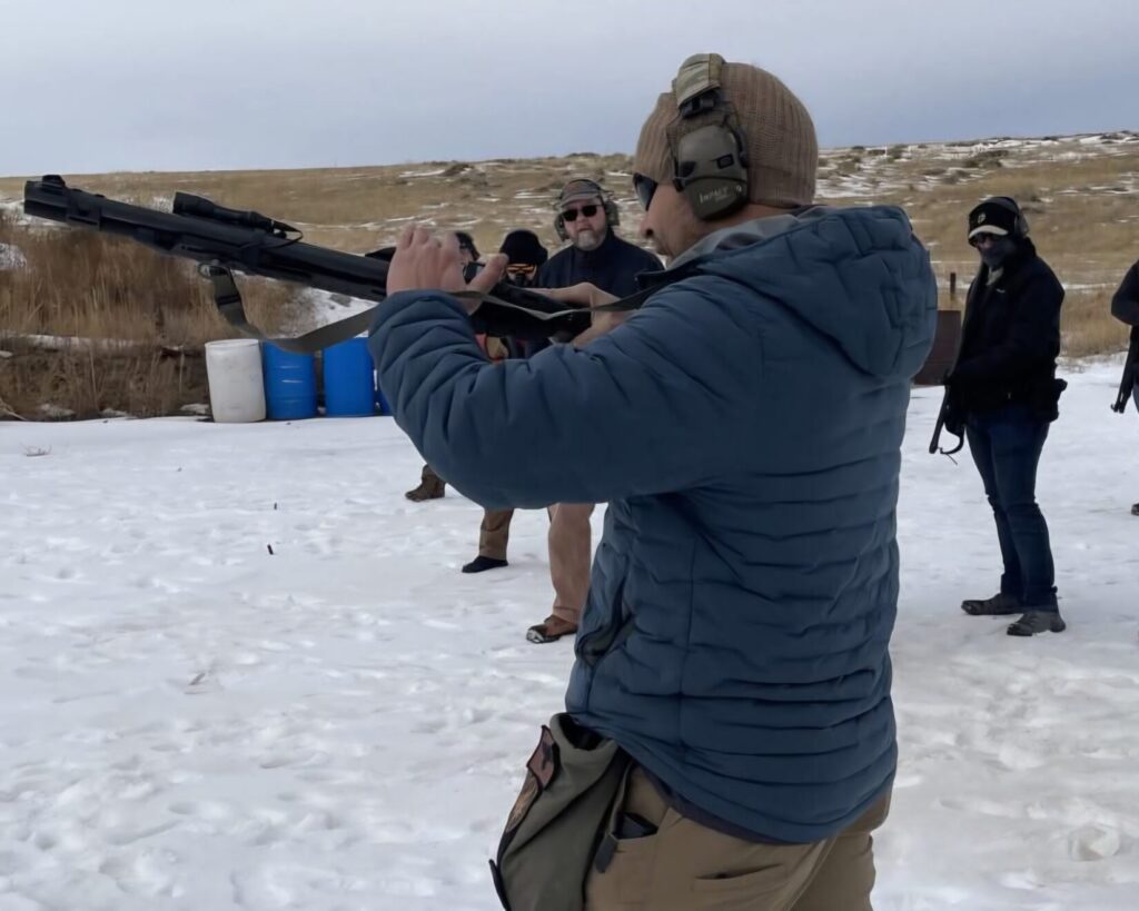 A man in a blue jacket standing in the snow with a shotgun while students watch
