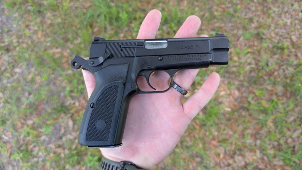 A person's hand holding a black semi-automatic pistol by the grip at an upward angle, showing the right side of the slide and frame against an outdoor, grassy background.