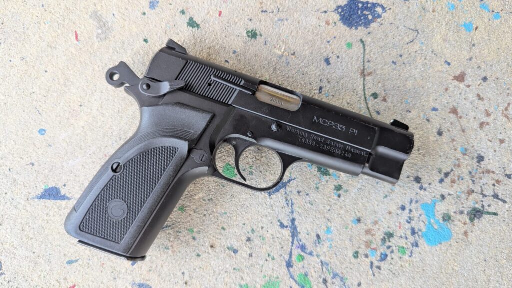 A top-down view of the left profile of a black semi-automatic pistol laying flat on a paint-splattered concrete surface, showing the safety lever, slide release, and "GIRSAN" engraving