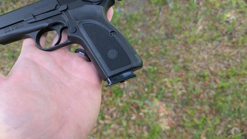 A black semi-automatic pistol held flat in an open palm, displaying the right-side profile of its slide, frame, and textured grip panels over a blurry ground background.