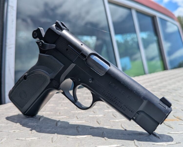 A black semi-automatic pistol resting upright on a silver diamond-plate surface, with the windows and side of a red vehicle in the background.