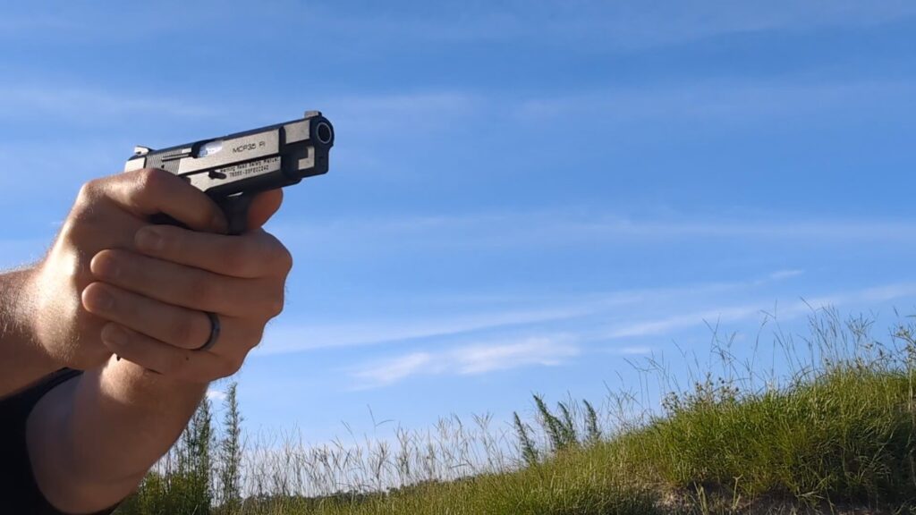 Side profile of a person's arms extended, holding a black handgun aimed straight ahead against a bright blue sky and grassy field background.