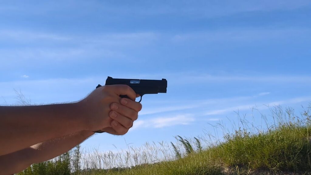 Close-up of a person's hands holding and aiming a black semi-automatic pistol against a bright blue sky with light clouds and a grassy hill in the lower right background.