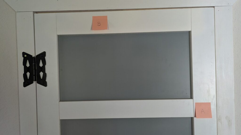 A close-up of a white door with frosted glass panels, featuring a pink sticky note labeled "B" near the top frame and another labeled "A" on the right frame.