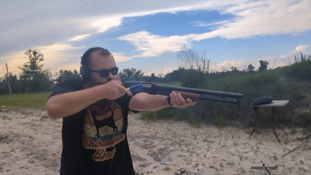 A man wearing sunglasses and ear protection aims a pump-action shotgun at an outdoor range while a blue shotgun shell ejects from the receiver.