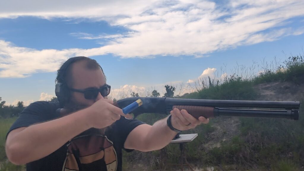 Close-up of a man in sunglasses and ear protection firing a pump-action shotgun, capturing a blue shell mid-ejection with faint smoke rising from the action.