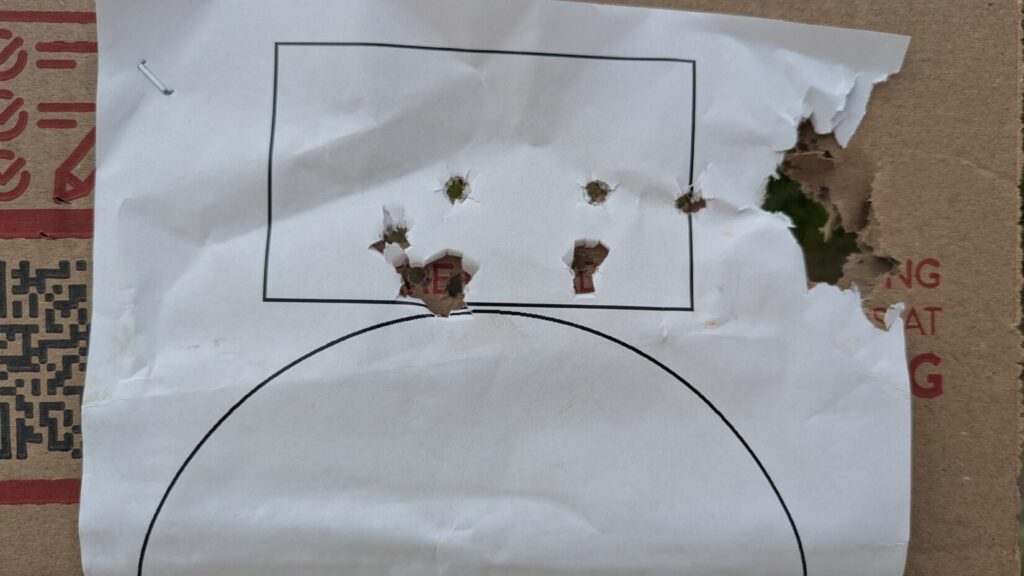 A close-up of a white paper target stapled to cardboard, showing several distinct bullet holes clustered around and inside a printed black rectangle.
