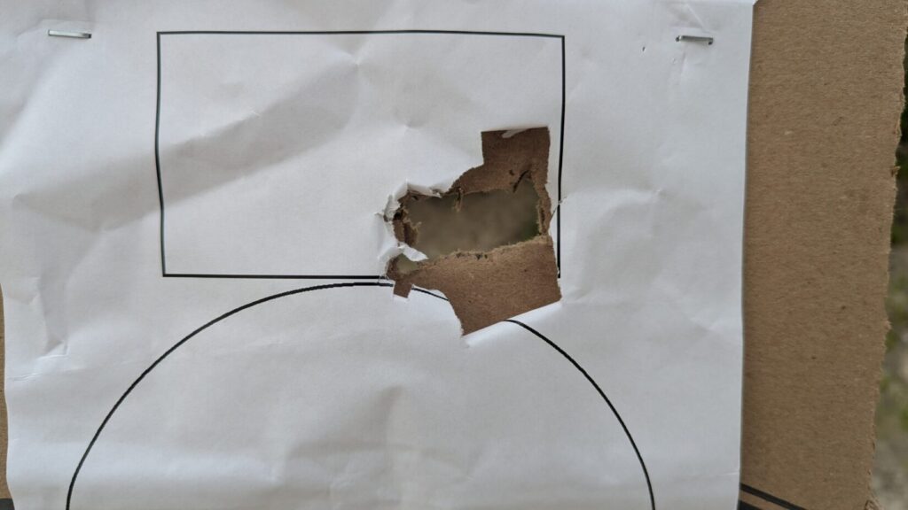 A white paper target on cardboard featuring a large, jagged hole blown through the right side of a printed black rectangle.