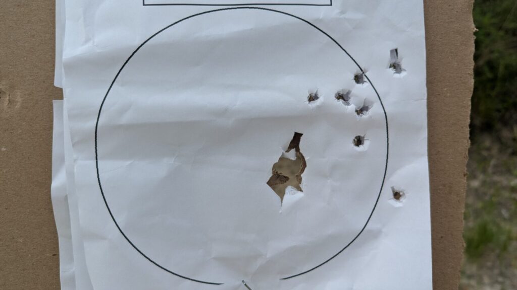 A white paper target showing a tight cluster of bullet impacts forming a large hole near the bottom right of a printed black circle, with a few scattered single holes nearby.