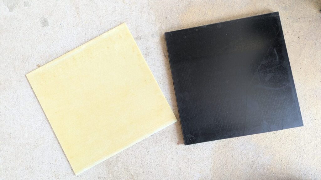 A top-down view of a square, light yellow fiberglass panel lying flat on concrete next to a separate square black composite panel.