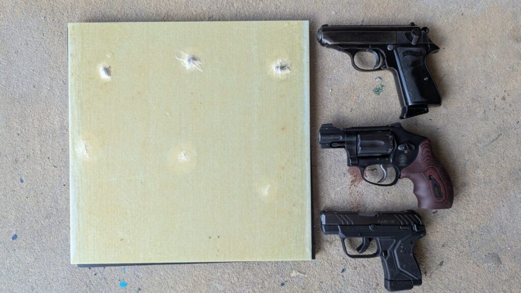 A top-down view showing a square, light yellow fiberglass panel with several impact marks on the left next to three compact handguns arranged vertically on a concrete surface.