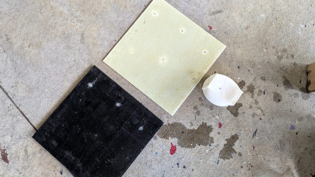 A flat, square tan tile on concrete showing several small impact craters, sitting next to a white molded plastic dome object.