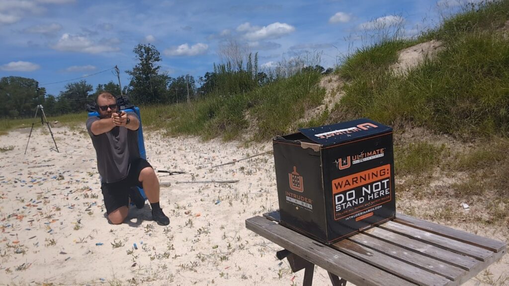 A kneeling man aims a small handgun toward an black "Intruder in a Box" cardboard target box sitting on a wooden bench at a sandy range.