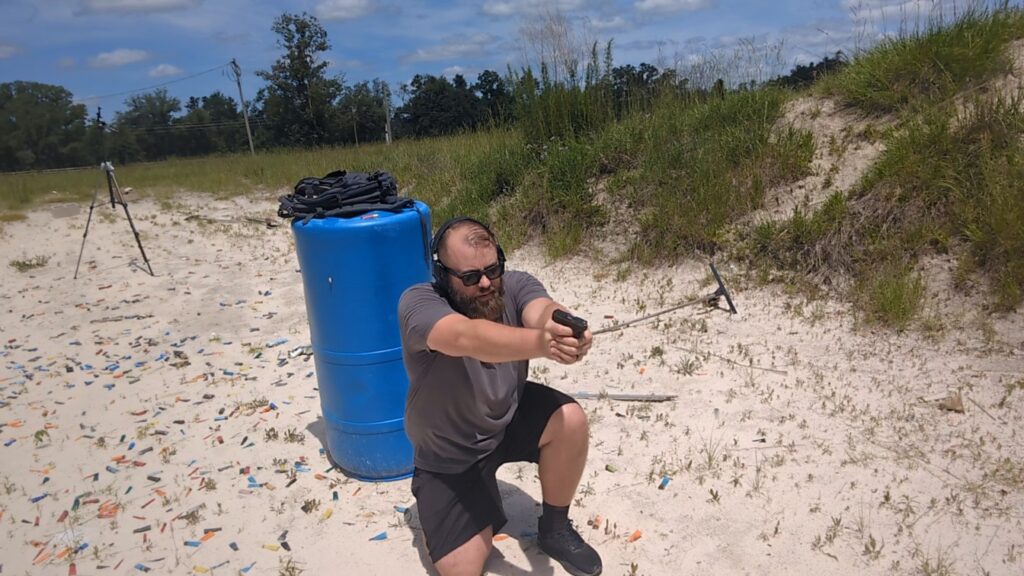 A man kneeling on a sandy outdoor range, aiming a small black semi-automatic pistol with both hands next to a blue plastic barrel.