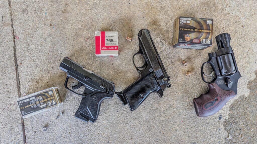 A top-down view of three compact handguns—two black semi-automatics and one black revolver with wood-grain grips—arranged on concrete next to boxes of Federal Premium and Remington ammunition.