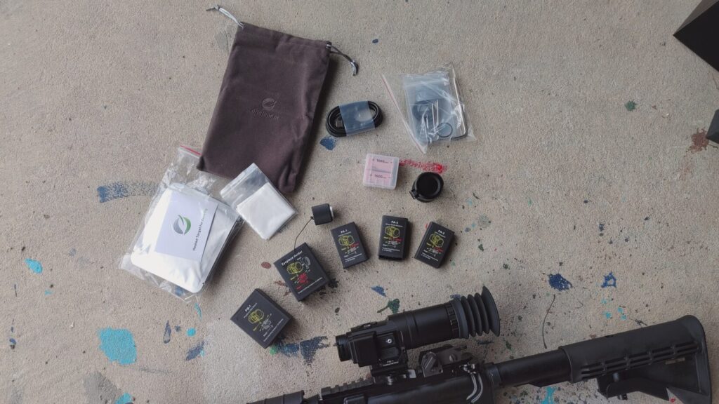 Overhead shot of the Mars Mini thermal scope disassembled on a concrete floor alongside its accessories, including batteries, cables, a storage pouch, and a lens cloth.