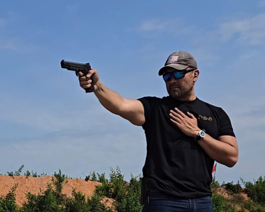 A man in a black shirt and ball cap aims a 1911 pistol at a range.