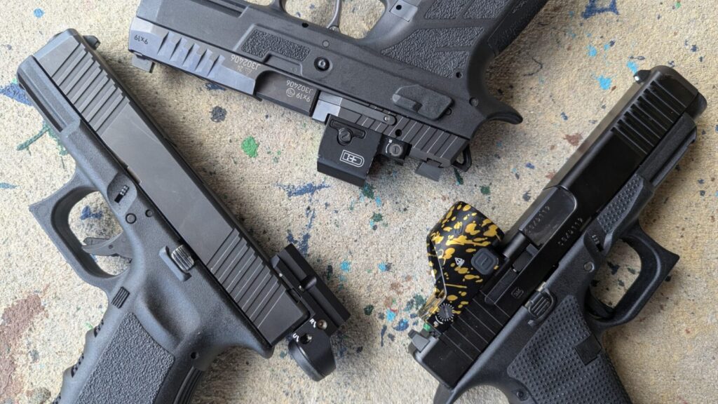 Overhead flat-lay of three handguns, highlighting the distinct footprint and lens sizes of a wide gold-splattered round optic, a square enclosed optic, and a slim tube-style micro sight.