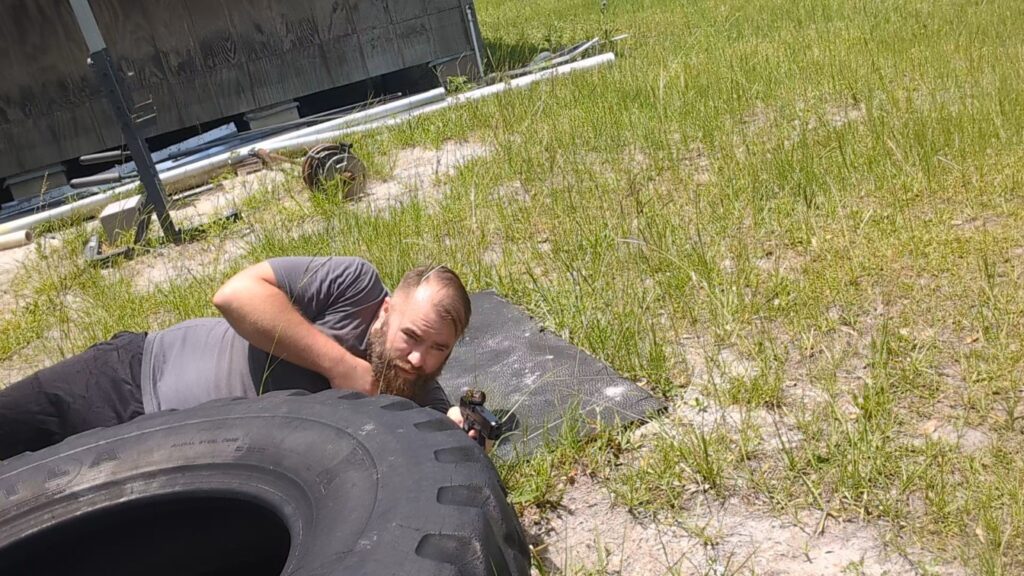 A man aiming a handgun prone from behind a large tire, utilizing a pistol mounted with a prominent, large-lens red dot sight.
