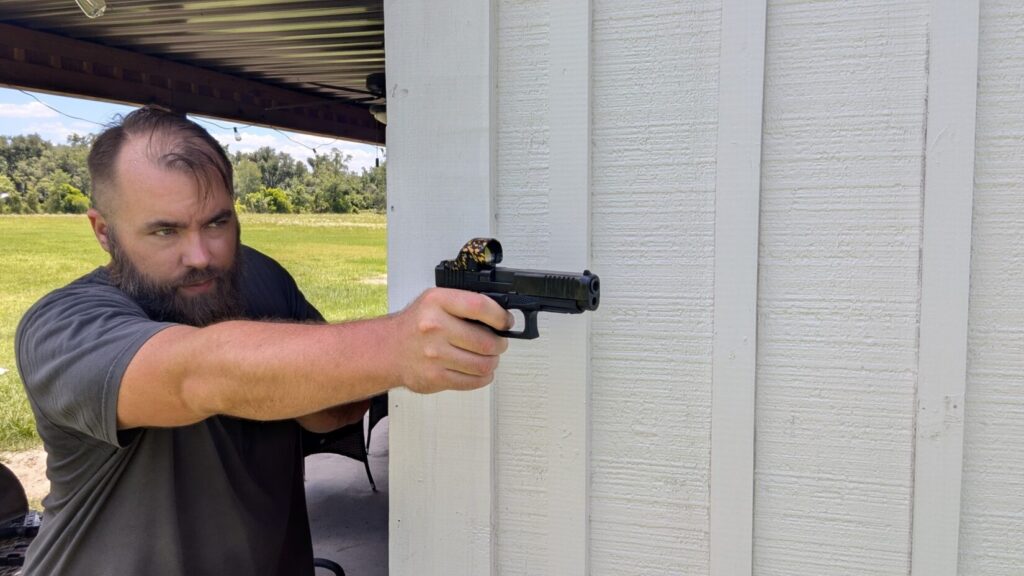 A man aiming a handgun from behind a white wall, showcasing the profile of a gold-splattered red dot sight with a large circular lens housing.