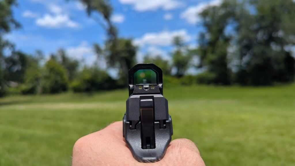 Point-of-view look through a compact enclosed pistol optic with a small, square-shaped lens window displaying a central aiming dot.